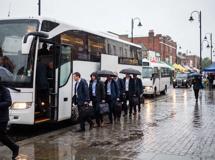 A white touring coach with a smaller minibus behind it at the kerb on Walthamstow High Street in steady rain, umbrellas up and headlights on, a corporate group with lanyards and laptop bags boarding beside the market stall canopies.